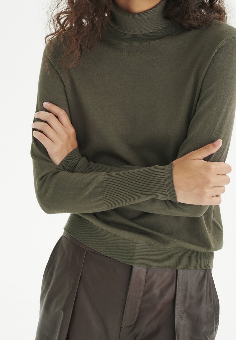 Pull à col roulé vert olive en tissu tricoté doux, avec poignets et ourlet côtelés. Porté avec un pantalon texturé marron foncé.