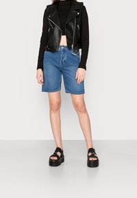 Černá kožená motorkářská vesta přes černý dlouhý rukáv, doplněná modrými džínovými šortkami a černými platformovými sandály.