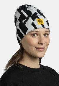 Knitted beanie in black and white with a geometric pattern, featuring a yellow Buff logo patch on the front. Soft texture, snug fit.