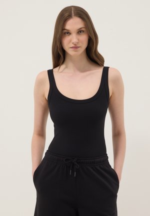 Jonge vrouw met lang bruin haar, gekleed in een zwart hemd en zwarte broek met koord, staand met handen in de zakken tegen een effen achtergrond.