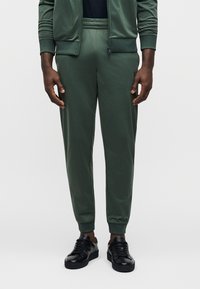 Πράσινο παντελόνι φόρμας με στενή γραμμή, ελαστική μέση και ριχτά μπατζάκια, κατασκευασμένο από λείο, ελαφρύ ύφασμα. Φοριέται με μαύρα αθλητικά παπούτσια.