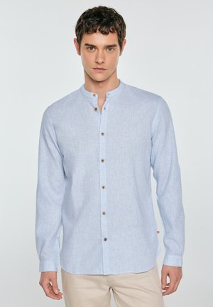 Młody mężczyzna w jasnoniebieskiej koszuli z kołnierzykiem typu mandarynka i beżowych spodniach, stojący na jednolitym jasnym tle.