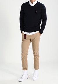 Uomo che indossa un maglione nero sopra una camicia bianca, pantaloni beige arrotolati, calzini bianchi e sneakers bianche, in piedi con le mani in tasca.