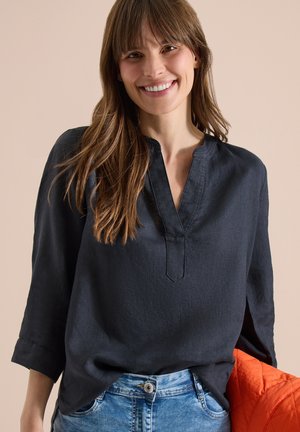 Femme souriante aux longs cheveux bruns, portant un haut noir à col en V et un jean bleu, tenant une veste matelassée orange sur un fond beige.