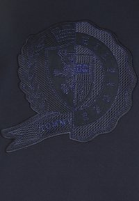 Spilla ricamata con un design a scudo in blu navy, con un leone e il testo "TOMMY H." circondato da una corona di alloro e elementi banner.