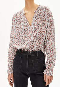 Kvinna som bär en blommig blus med lång ärm, V-ringning och dragsko-detalj, instoppad i höga svarta jeans med framfickor.