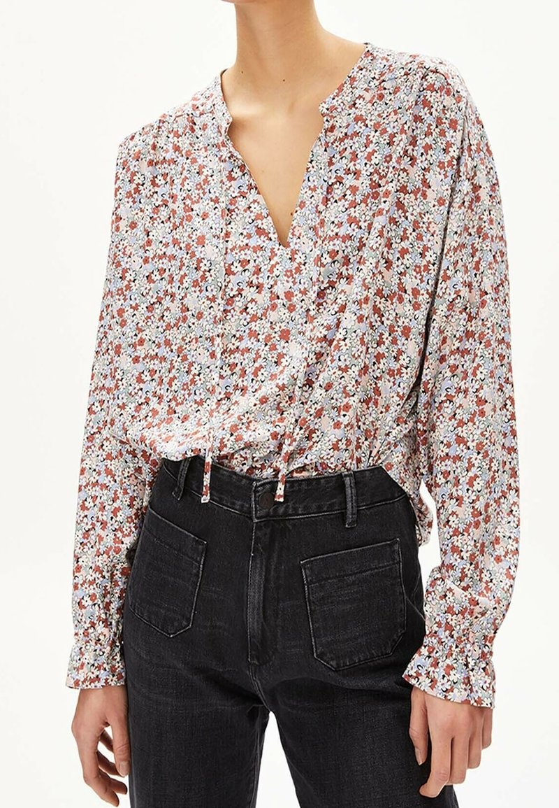 Kvinna som bär en blommig blus med lång ärm, V-ringning och dragsko-detalj, instoppad i höga svarta jeans med framfickor.