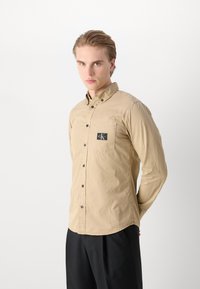Camicia beige con bottoni realizzata in tessuto leggero. Presenta una tasca sul petto con una toppa nera e bottoni scuri. Texture liscia, design aderente.