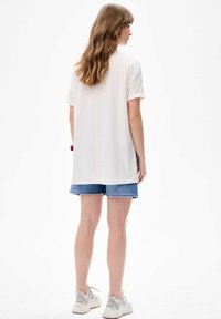 Weißes Kurzarmhemd mit lockerer Passform, kombiniert mit Jeansshorts. Das Outfit zeichnet sich durch ein schlichtes Design und einen leichten, atmungsaktiven Stoff aus.