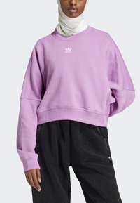 Levandulová skrátená mikina s pruhovaným výstrihom a nadmernými rukávmi, so malým bielym logom Adidas na hrudi. Čierne štruktúrované nohavice.