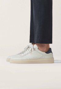 Zapatillas de cuero blancas con una suela color crema, con un detalle en azul marino en el talón, diseño de cordones y detalles perforados en los laterales.