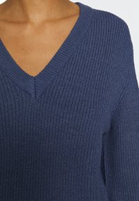 Pull en maille bleu marine avec une texture côtelée, col en V et manches longues, présentant des coutures soignées et une coupe décontractée.