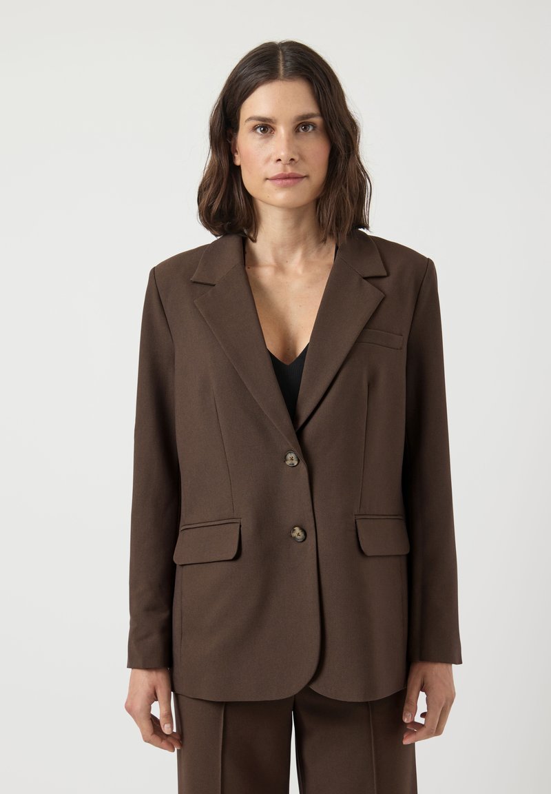 Femme aux cheveux foncés mi-longs, portant un blazer marron à deux boutons et un pantalon assorti, debout devant un fond clair uni.