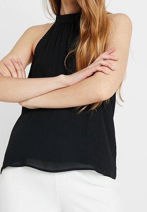 Frau mit langen Haaren, die ein ärmelloses schwarzes Neckholder-Top und weiße Hosen trägt, Arme vor der Brust verschränkt.