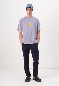 Lilla kortermet t-skjorte med en gul "GUESS" logo, kombinert med marineblå bukser og svarte sandaler, toppet med en lyseblå caps.