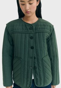 Giacca verde trapuntata con collo rotondo, quattro bottoni e due tasche frontali inclinate. Presenta cuciture verticali per texture.