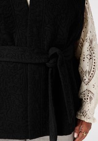 Zwarte gewatteerde vest met een strikceintuur, voorzien van een gestructureerd oppervlak. De binnenkant toont een roomkleurige kantensleeve met intrigerende patronen.