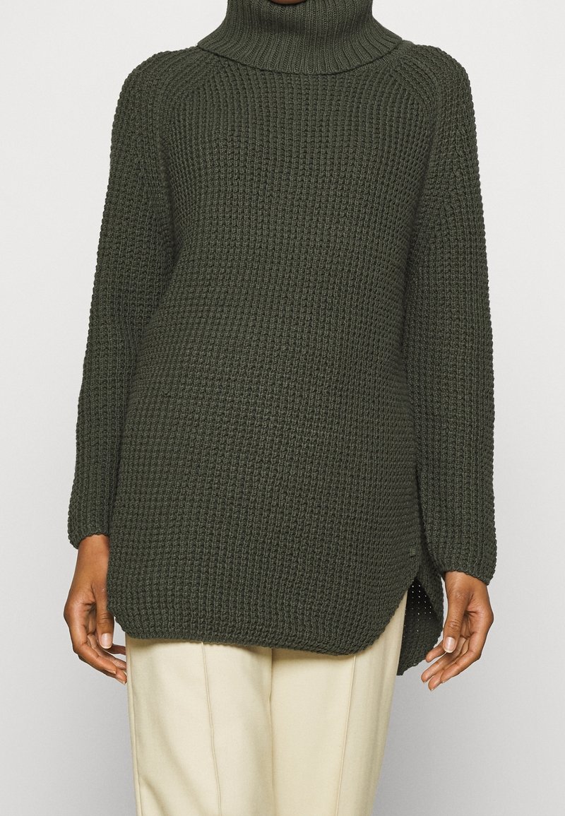 Pull à col roulé en maille épaisse vert foncé à manches longues porté sur un pantalon beige, avec une fente latérale et un motif texturé.