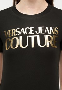 Μαύρο T-shirt με λαμπερό χρυσό κείμενο "Versace Jeans Couture" που φοριέται από άτομο, ορατό από τον λαιμό μέχρι το στήθος.