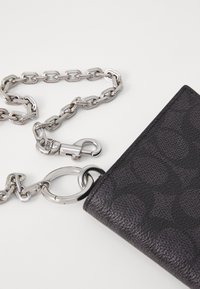 Portefeuille noir texturé avec une chaîne en argent, doté d'un fermoir et d'un attache en anneau circulaire. Motif gaufré remarquable sur la surface.