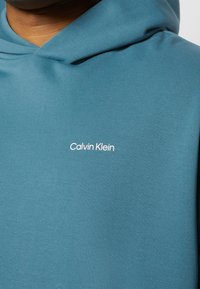 Sudadera con capucha azul con pequeño logo blanco de "Calvin Klein" centrado en el pecho, usada por una persona de piel oscura.