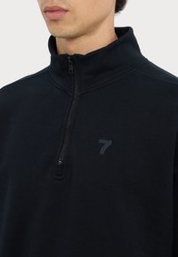 Pulover negru cu guler înalt și fermoar pe un sfert, realizat dintr-un material moale. Prezintă un mic logo gri închis "7" pe pieptul stâng.