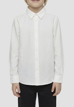 Dijete odjeveno u bijelu košulju s dugim rukavima i crne hlače, stoji ispred jednobojne pozadine.