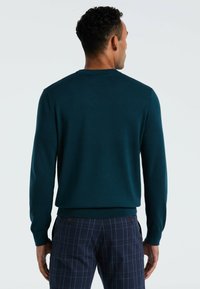 Pull en maille teal avec encolure ras du cou et manches longues. Texture lisse, silhouette ajustée, associé à un pantalon à carreaux marine.