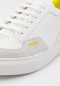 Witte leren sneaker met grijze suède accenten, geperforeerde neus en witte veters. Voorzien van een geel "HUGO" logo en een gestructureerde rubberen zool.