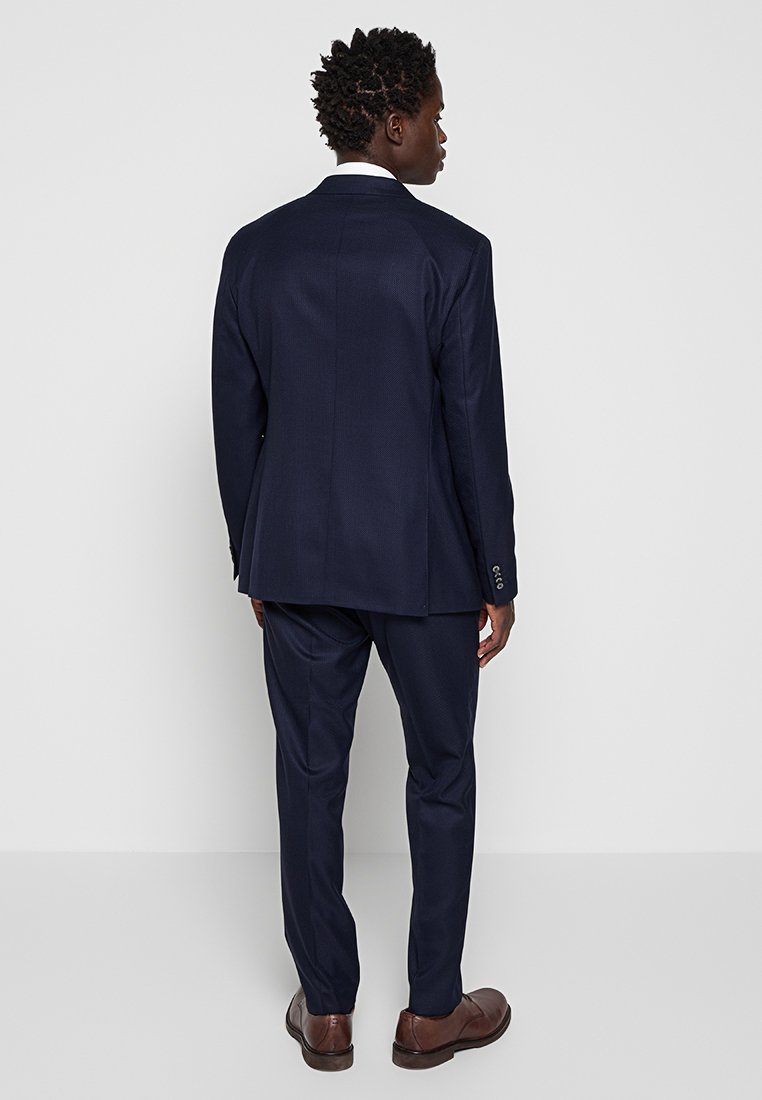 Marineblauer maßgeschneiderter Anzug mit strukturierter Oberfläche, ausgestattet mit Jackett mit einem Schlitz und schmal geschnittenen Hosen, ergänzt durch braune Anzugschuhe.