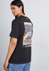 Sort bomulds t-shirt med et stort grafik af en Porsche bil og tekst på ryggen. Korte ærmer, rund hals, afslappet pasform.