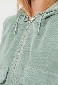 Πράσινο σακάκι από κορντόρι με φερμουάρ, κουκούλα με κορδόνι και δύο μπροστινές τσέπες. Υφασμάτινη υφή με ριγερό μοτίβο.