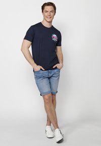 Tums zila kokvilnas T-kreklu ar grafiku uz kreisā krūts, kombinēts ar gaišiem džinsu šortiem un baltām sporta kedām. Modelis pozē ikdienišķi.