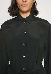 Femme portant une robe-chemise noire à boutons, avec un col pointu et une ceinture à la taille, montrée du cou aux hanches sur fond uni.