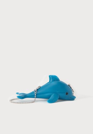 Pochette bleue en forme de dauphin avec bandoulière chaîne argentée posée sur une surface blanche, avec un petit détail œil noir et le nom de la marque sur la nageoire.