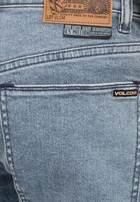 Jeans en denim bleu clair avec une patch en cuir portant le logo et le texte, des détails de couture, et une étiquette de marque noire sur la poche arrière.