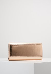 Bolso de mano rectangular metálico en color rosa oro con una superficie lisa y brillante sobre un pedestal blanco de exhibición.