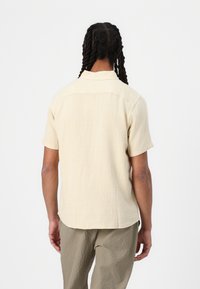 Kort geblokte shirt in lichtbeige met een gestructureerde afwerking, voorzien van een standaard kraag en een rechte zoom. De achterkant toont subtiele verticale lijnen.