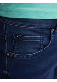 Jeans blu scuro con tasca posteriore con cuciture visibili, un piccolo bottone e una finitura in tessuto testurizzato. Il bordo superiore mostra una camicia verde.