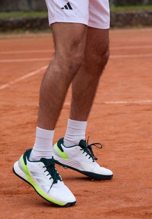 Nogi osoby ubranej w białe szorty Adidas, białe skarpetki i białe tenisówki z czarnymi i neonowo żółtymi detalami na ceglanej nawierzchni kortu tenisowego.