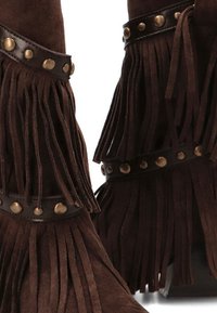 Botas de ante marrón con detalles de flecos, adornadas con tachuelas de bronce y una correa de cuero suave, que muestran un diseño afilado.