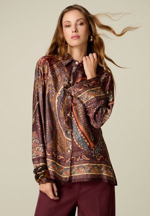 Junge Frau mit langen Haaren trägt ein gemustertes Hemd mit Paisley-Motiven und bordeauxrote Hose, posiert mit einer Hand am Hals.