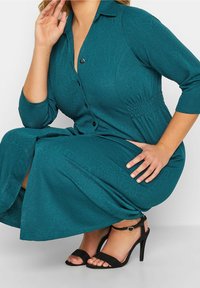 Turkusowa sukienka z rękawami do łokcia, teksturowanym wzorem, zapięciem na guziki z przodu, dopasowaną talią i rozkloszowanym dołem. Noszona z czarnymi sandałkami na cienkich paskach.