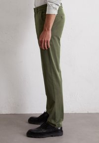 Oliwkowe spodnie w dopasowanym kroju o gładkiej teksturze, z bocznymi kieszeniami i subtelnym detalem na tylnej kieszeni, w zestawieniu z czarnymi butami.