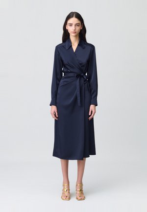Robe portefeuille en satin bleu marine avec des manches longues, un col, une ceinture à la taille et une longueur midi. Accompagnée de sandales dorées à lanières.