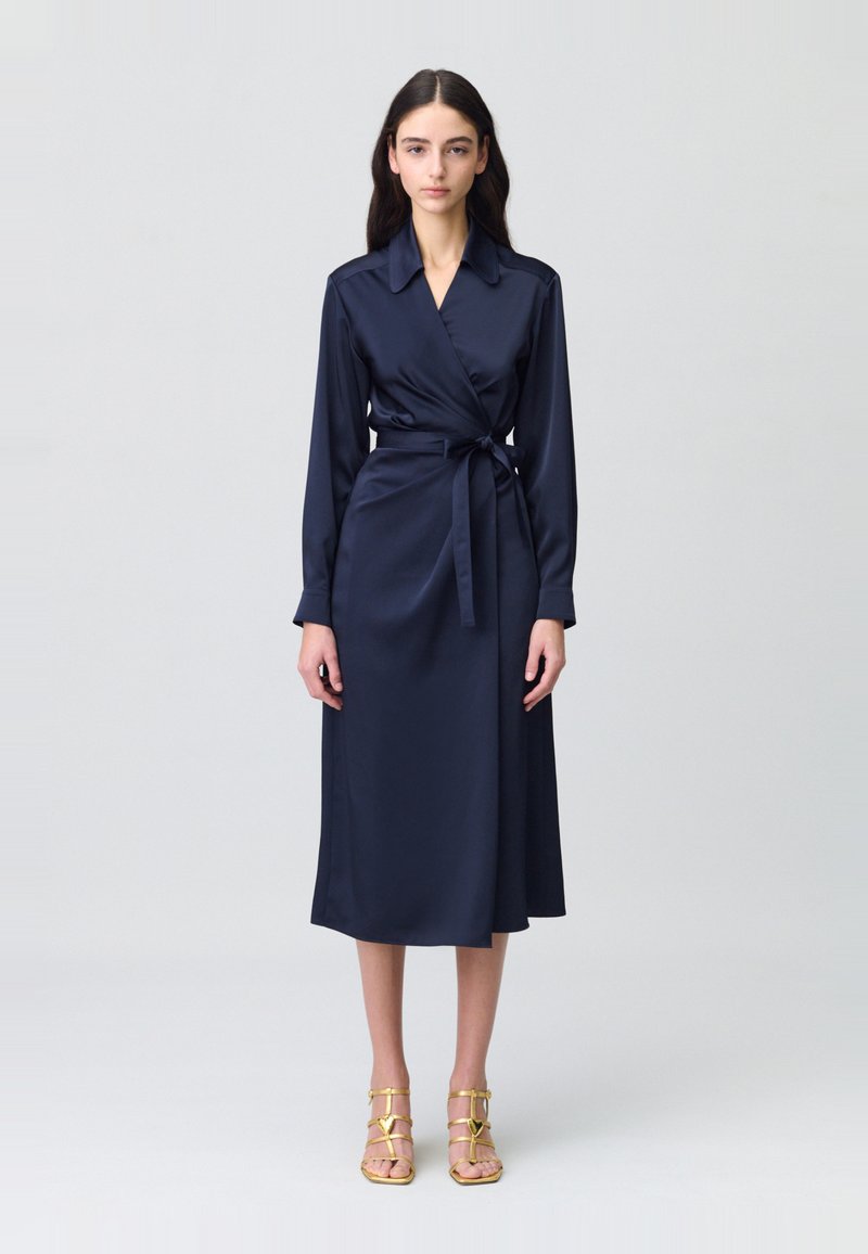 Vestido envolvente de satén azul marino con mangas largas, cuello, cinturón en la cintura y una longitud midi. Acompañado de sandalias doradas con tiras.