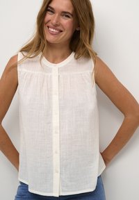 Blouse sans manches beige clair en tissu texturé, avec un devant boutonné et un col rond. Design simple et décontracté.