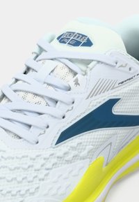 Λευκό αθλητικό παπούτσι με επάνω μέρος από διχτυωτό υλικό, μπλε λεπτομέρειες και κίτρινη σόλα. Διαθέτει υφές και έντονο λογότυπο στη γλώσσα.