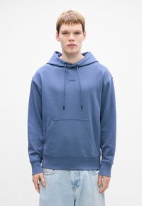 Giovane uomo che indossa un hoodie blu di BOSS con lacci e tasca frontale, abbinato a jeans azzurri, in piedi contro uno sfondo bianco.