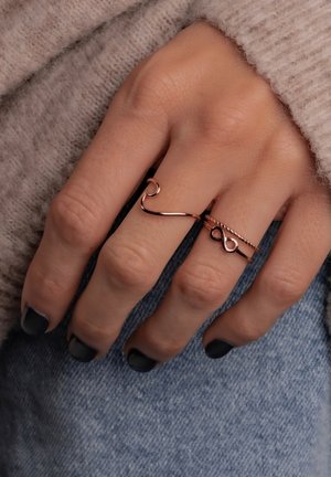 Due anelli in oro rosa su una mano: uno ha un design ondulato, l'altro ha una bandarica attorcigliata con un simbolo dell'infinito; lo smalto nero contrasta.
